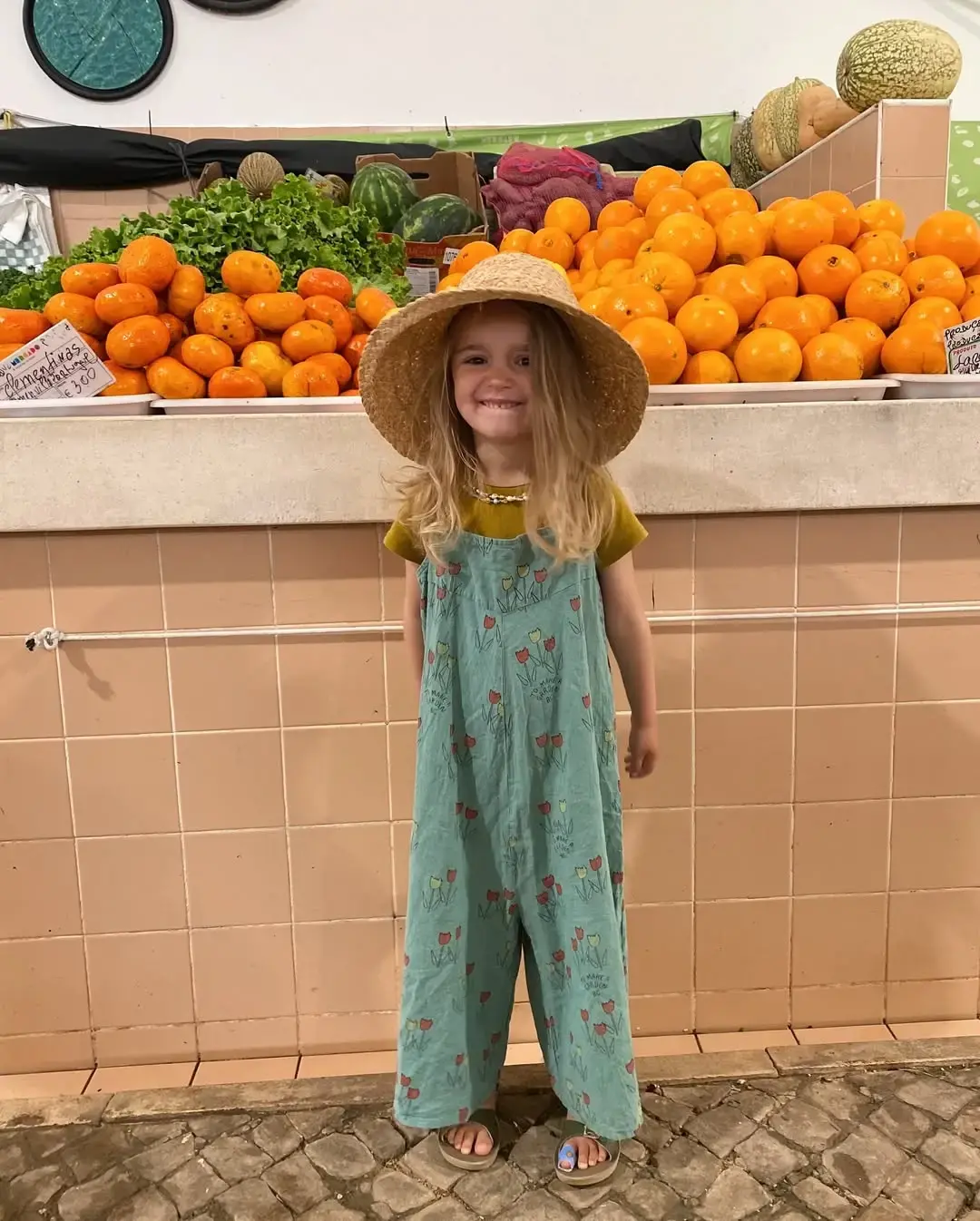 suki criança menina com macacão verde e um chapéu de palha no mercado junto às laranjas e tomates e melão